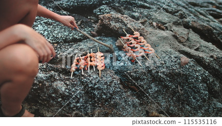 A person is sitting on a large rock while holding and eating food skewered on sticks, likely a seafood barbecue dish. The individual appears relaxed and enjoying the outdoor dining experience. A person is sitting on a large rock while holding and eating food skewered on sticks, likely a seafood barbecue dish. The individual appears relaxed and enjoying the outdoor dining experience. 115535116