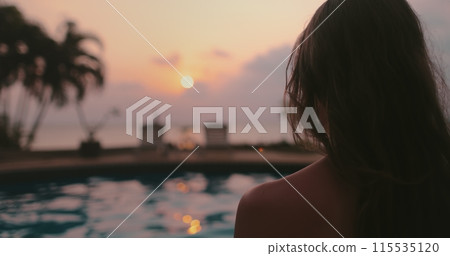 Woman relaxes near swimming pool at sunset. Long hair brunette girl back view enjoys colorful sky reflected in water, palm trees in background. Tropical resort, travel, exotic vacation in Thailand Woman relaxes near swimming pool at sunset. Long hair brunette girl back view enjoys colorful sky reflected in water, palm trees in background. Tropical resort, travel, exotic vacation in Thailand 115535120