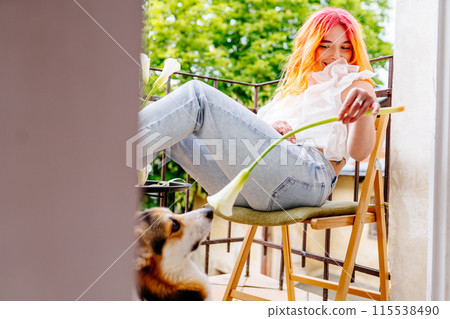 Playful woman with pink hair is sitting on a chair with a flower in her hand. A dog is standing behind her, looking at her. The scene is peaceful and relaxing, female enjoying time outdoors Playful woman with pink hair is sitting on a chair with a flower in her hand. A dog is standing behind her, looking at her. The scene is peaceful and relaxing, female enjoying time outdoors 115538490