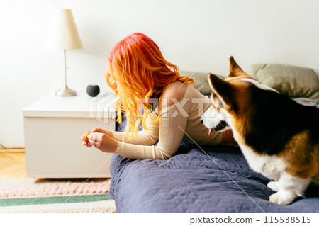 Thoughtful sad woman with bright orange hair lying with dog on couch at home. 115538515