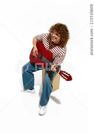 Isometric view. Young smiling curly-haired man in casual clothes siting on wooden box and playing guitar isolated on white background Isometric view. Young smiling curly-haired man in casual clothes siting on wooden box and playing guitar isolated on white background 115538609