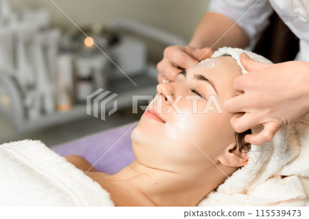 Young woman enjoying facial massage, lying on spa bed indoor at beauty salon. 115539473