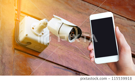 A hand holding a mobile phone is placed on a picture of a CCTV camera at a home. A hand holding a mobile phone is placed on a picture of a CCTV camera at a home. 115539918