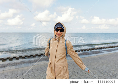 Smiling happy woman walking by Nord sea coast wear beige coat  sunglasses enjoys windy day cloudy sky background. Female person have fun hiking adventure travel at Baltic sea seaside 115540091