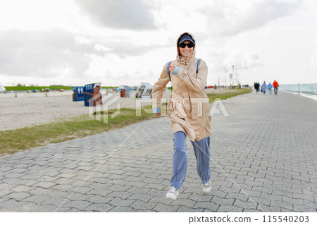 Smiling happy woman walking by Nord sea coast wear beige coat  sunglasses enjoys windy day cloudy sky background. Female person have fun hiking adventure travel at Baltic sea seaside 115540203