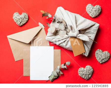 Zero waste Valentine's Day concept and mock up on red. Eco-friendly gift cloth wrapping in Furoshiki style, craft paper envelope,empty greetings card. Top down view or flat lay. Copy space for design 115541693