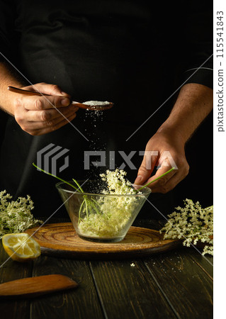 Adding sugar to a bowl of elderberries before making kvass. A spoon in the hands of a herbalist above a kitchen table with medicinal herbs. Space for advertising 115541843
