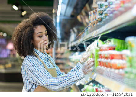 young African American woman in a work apron and disposable gloves works in a supermarket, stands among the shelves and lays out products and merchandise. young African American woman in a work apron and disposable gloves works in a supermarket, stands among the shelves and lays out products and merchandise. 115542031