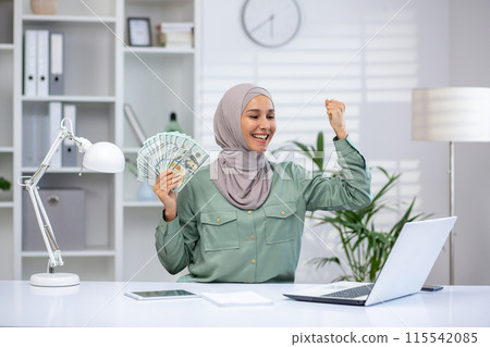 Happy young muslim businesswoman in hijab sitting in office at work desk. holds a fan of banknotes in his hands and rejoices in financial success. 115542085