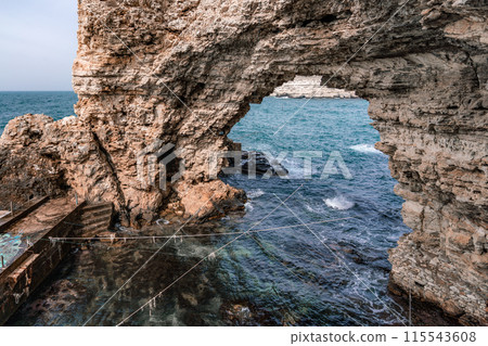A large rock archway with a small pool of water in front of it. The water is calm and the sky is clear. A large rock archway with a small pool of water in front of it. The water is calm and the sky is clear. 115543608