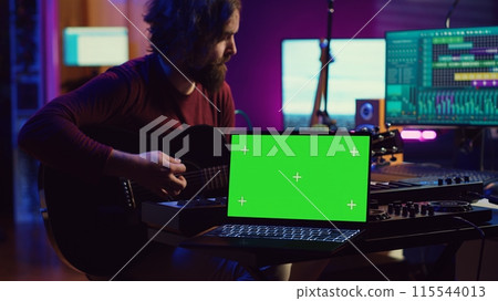 Guitarist playing his acoustic instrument next to greenscreen on laptop, learning new strings accords to play on guitar in home studio. Musician artist developing his singing skills. Camera A. 115544013