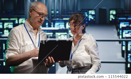 Adept mechanics looking around high tech data center, using laptop to crosscheck disaster recovery plan and assess server clusters in need of replacement, preventing pitfalls 115544022