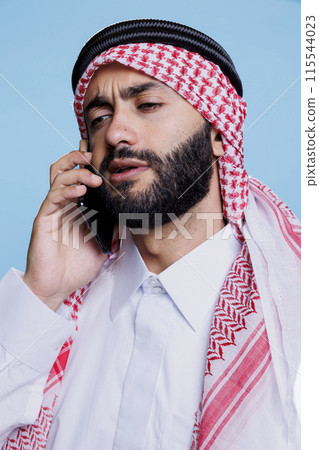 Muslim man wearing traditional headscarf speaking on smartphone with confused expression. Young arab person dressed in red and white checkered ghutra speaking on mobile phone Muslim man wearing traditional headscarf speaking on smartphone with confused expression. Young arab person dressed in red and white checkered ghutra speaking on mobile phone 115544023