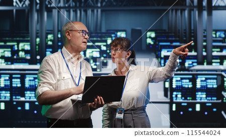 Proficient repairmen looking around high tech data center, using laptop to crosscheck disaster recovery plan and assess server rows in need of replacement, preventing hindrances 115544024
