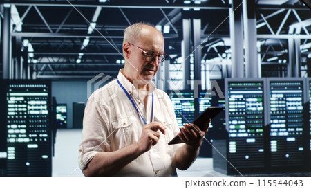 Octogenarian professional navigating network of server rigs in industrial base of operations. Admin with tablet doing review and optimizations in high tech data center, preventing system flaws 115544043