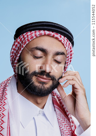 Smiling muslim man with closed eyes wearing ghutra headscarf dreaming closeup. Arab person in checkered red and white headdress feeling sleepy and daydreaming on blue background Smiling muslim man with closed eyes wearing ghutra headscarf dreaming closeup. Arab person in checkered red and white headdress feeling sleepy and daydreaming on blue background 115544052