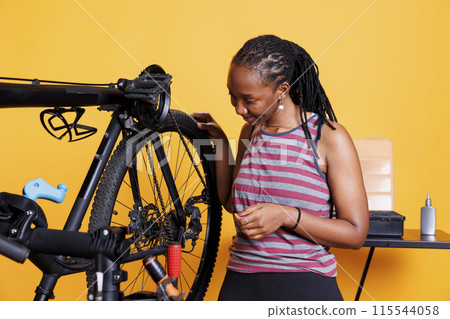 Against an isolated background, sports-loving lady performs yearly maintenance and adjustment of bicycle tyre. Youthful african american woman inspects her bike wheel for leisure cycling. 115544058