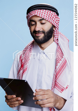 Muslim man wearing traditional clothes holding digital tablet and exploring content. Smiling arab dressed in islamic white thobe and ghutra headscarf scrolling social media on touchscreen Muslim man wearing traditional clothes holding digital tablet and exploring content. Smiling arab dressed in islamic white thobe and ghutra headscarf scrolling social media on touchscreen 115544062