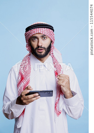Arab man enjoying online game on smartphone and looking at camera with surprised expression. Muslim player engaging in online entertainment and showing victory with clenched fist Arab man enjoying online game on smartphone and looking at camera with surprised expression. Muslim player engaging in online entertainment and showing victory with clenched fist 115544074