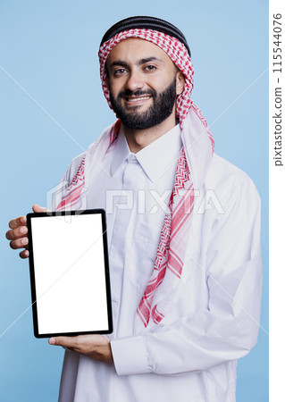 Smiling muslim man dressed in traditional thobe confidently showcasing digital tablet with blank screen for copy space. Cheerful arab person showing white touchscreen and looking at camera 115544076