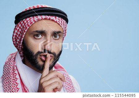 Man dressed in muslim clothes asking to keep secret while holding finger on lips and looking at camera. Arab person wearing headscarf with rope band showing silent gesture portrait Man dressed in muslim clothes asking to keep secret while holding finger on lips and looking at camera. Arab person wearing headscarf with rope band showing silent gesture portrait 115544095