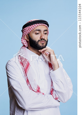 Pensive man wearing traditional muslim clothes rubbing chin and standing in contemplation pose in studio. Puzzled arab person dressed in ghutra and thobe thinking and looking away 115544104