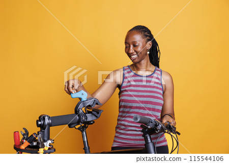 Healthy sports-loving black woman inspects damaged bicycle against isolated yellow background. Active lady carefully and efficiently setting up repair station for bike maintenance and adjustments. 115544106