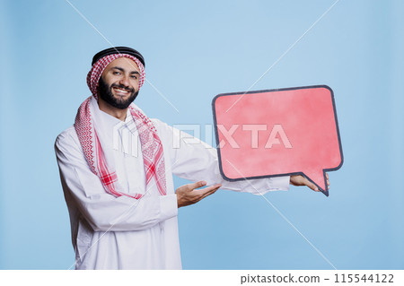 Cheerful man dressed in muslim traditional clothes showing at blank red dialog banner and looking at camera. Smiling carefree arab holding speech bubble mockup with copy space Cheerful man dressed in muslim traditional clothes showing at blank red dialog banner and looking at camera. Smiling carefree arab holding speech bubble mockup with copy space 115544122