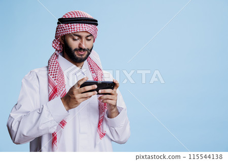 Smiling muslim man dressed in traditional islamic clothes playing online game on smartphone. Arab person enjoying virtual entertainment with mobile phone software app in studio 115544138