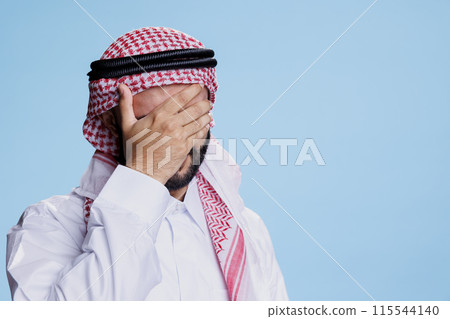 Man wearing muslim ghutra headdress holding forehead while suffering from migraine. Arab person touching head in pain and having headache while posing in studio on blue background Man wearing muslim ghutra headdress holding forehead while suffering from migraine. Arab person touching head in pain and having headache while posing in studio on blue background 115544140