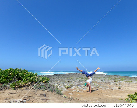 Man does split leg Handstand on coral rocks on the beach as wave crash in the distance Man does split leg Handstand on coral rocks on the beach as wave crash in the distance 115544346