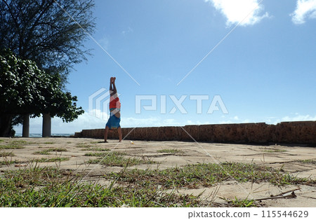 Man Handstanding stone foundation along cliff shore next to shallow ocean Man Handstanding stone foundation along cliff shore next to shallow ocean 115544629