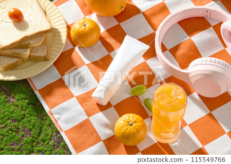 Flat lay cosmetic tube in white color on orange picnic mat with checkered pattern, next to a glass of orange juice, fresh orange, a dish of sandwich and a headphone. Blank space for text, top view 115549766