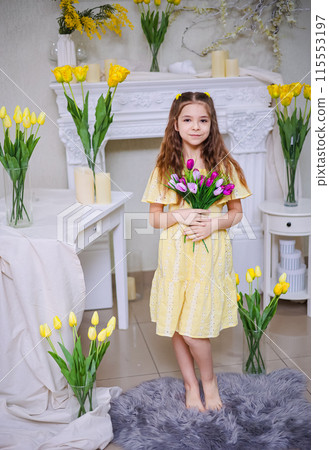 Cute girl in a yellow dress with a bouquet of purple flowers on a background of yellow tulips. Congratulations on Mother's Day, Women's Day, March 8th. Cute girl in a yellow dress with a bouquet of purple flowers on a background of yellow tulips. Congratulations on Mother's Day, Women's Day, March 8th. 115553197
