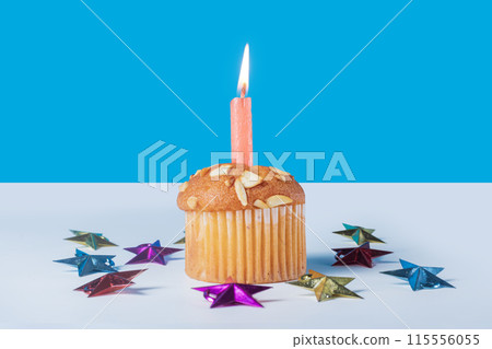 Birthday cupcake with glowing candle and colorful stars around it on white table with blue background. Closeup Birthday cupcake with glowing candle and colorful stars around it on white table with blue background. Closeup 115556055