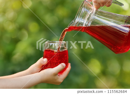 child hands holding glass and cherry juice pouring from jug with green garden on the background child hands holding glass and cherry juice pouring from jug with green garden on the background 115562684