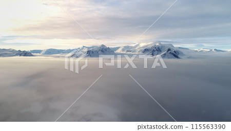 Majestic snow-capped mountain peaks above sunset clouds. Fly over morning fog cloudscape in Antarctica. Ocean aerial view surface of Antarctic continent. Wild nature background. Drone flight Majestic snow-capped mountain peaks above sunset clouds. Fly over morning fog cloudscape in Antarctica. Ocean aerial view surface of Antarctic continent. Wild nature background. Drone flight 115563390