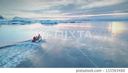 Antarctica travel and exploration, people riding motor boat. Zodiac boat sailing cold polar ocean water surface in sunset light. Discover the beauty of South Pole. Aerial winter landscape, drone 115563480