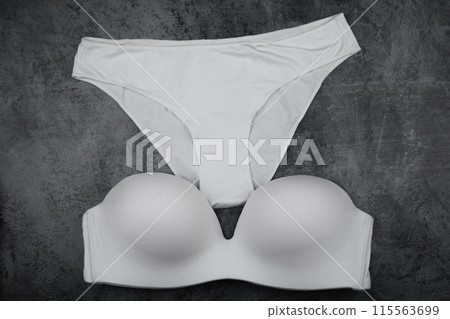 A white bra with stiff cups and a push-up made of white smooth satin fabric lies on a gray background, white ceta panties lie above the bra. A set of underwear. High quality photo A white bra with stiff cups and a push-up made of white smooth satin fabric lies on a gray background, white ceta panties lie above the bra. A set of underwear. High quality photo 115563699