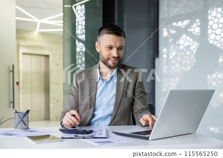 Smiling businessman working on laptop in modern office, analyzing documents and taking notes. Professional and focused in corporate environment. Smiling businessman working on laptop in modern office, analyzing documents and taking notes. Professional and focused in corporate environment. 115565250