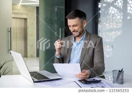 Smiling businessman working on laptop, analyzing documents in a modern office setting. Celebrating success and achievement. Smiling businessman working on laptop, analyzing documents in a modern office setting. Celebrating success and achievement. 115565253