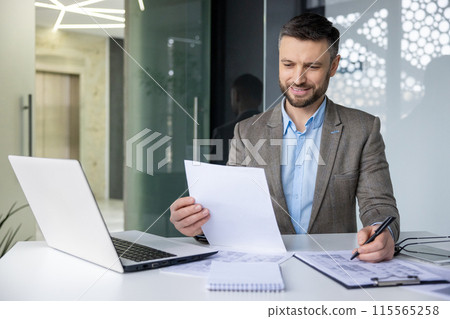 Smiling businessman working at desk with laptop, reviewing documents and taking notes in a modern office Smiling businessman working at desk with laptop, reviewing documents and taking notes in a modern office 115565258
