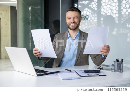 Confident smiling businessman showcasing documents at his desk in a modern office setting. Professional workspace with laptop and office supplies. Confident smiling businessman showcasing documents at his desk in a modern office setting. Professional workspace with laptop and office supplies. 115565259