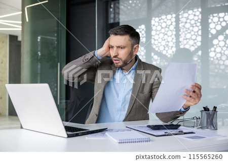 Stressed businessman holding documents and working on a laptop in a modern office. Man is facing difficulty while analyzing data or dealing with financial problems. 115565260