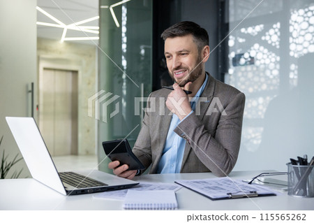 Smiling businessman using laptop and tablet while working at his desk in a modern office environment. Business, technology, and professionalism concepts. Smiling businessman using laptop and tablet while working at his desk in a modern office environment. Business, technology, and professionalism concepts. 115565262