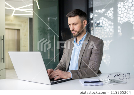Professional businessman working on a laptop in a modern office setting, focused on his tasks. Background features glass walls and a notebook on the desk. 115565264