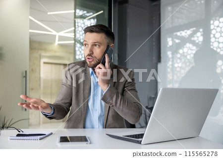 Businessman in a suit speaking on a phone call and appearing confused while sitting in a modern office ,with a laptop and tablet on the table. 115565278