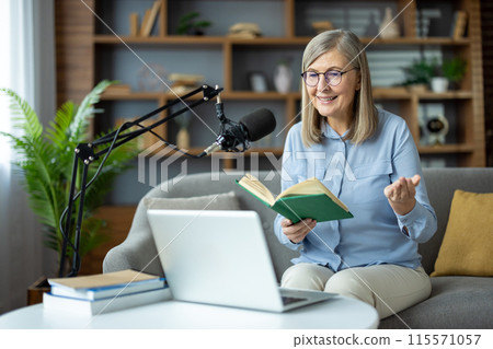 Senior woman recording podcast at home, holding a book and smiling, seated in front of a laptop and microphone. 115571057