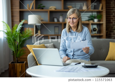 Senior woman working from home with a laptop and documents, sitting on a cozy sofa in a stylish living room, productive and content. 115571063