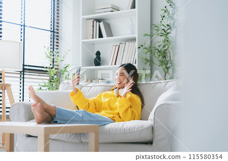 Happy young asian woman  wearing headset and relaxing on comfortable couch at home 115580354